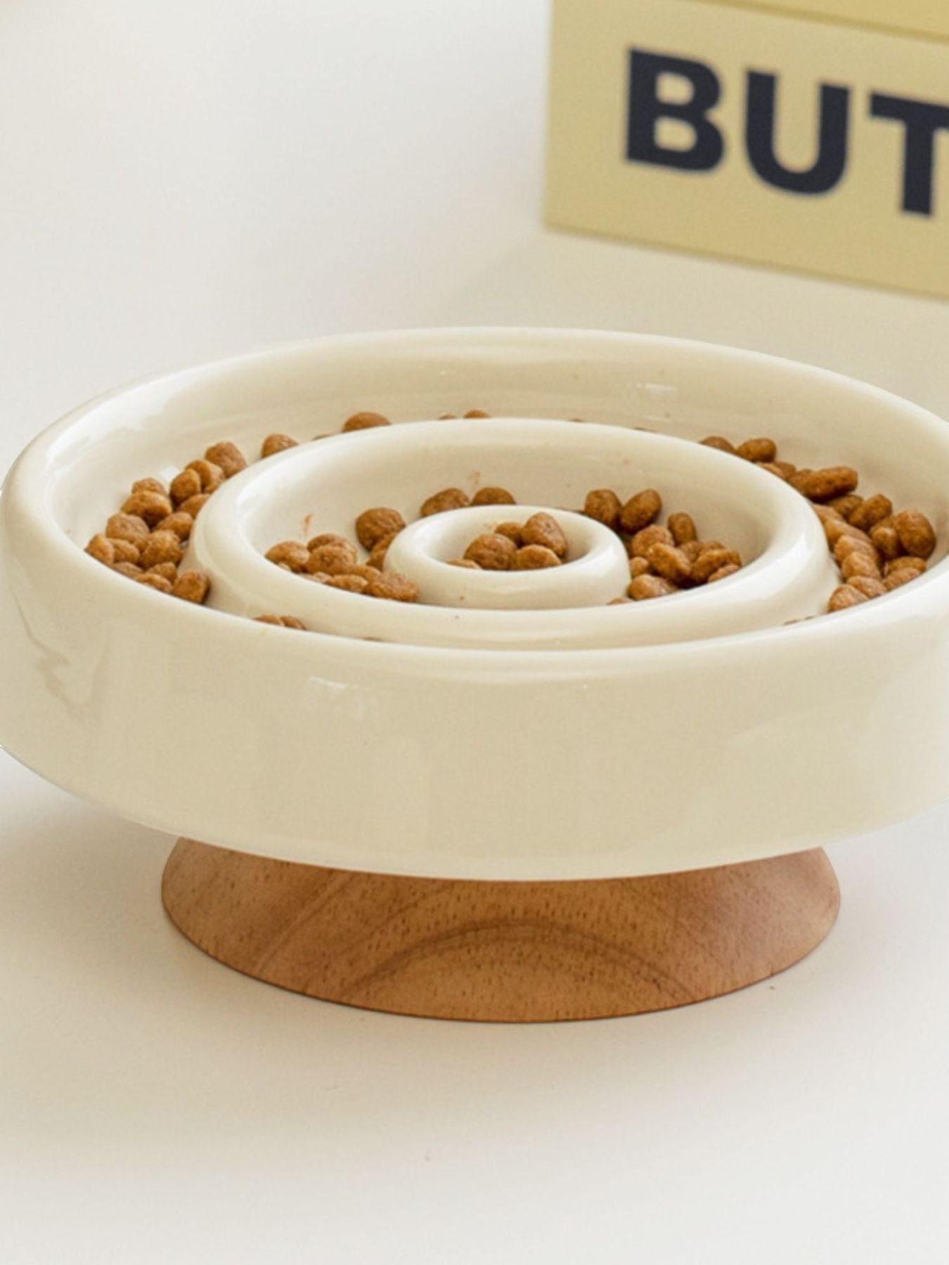 Little Pets Club white ceramic slow feeder dog and cat bowl with spiral pattern on a wooden base, filled with kibble.