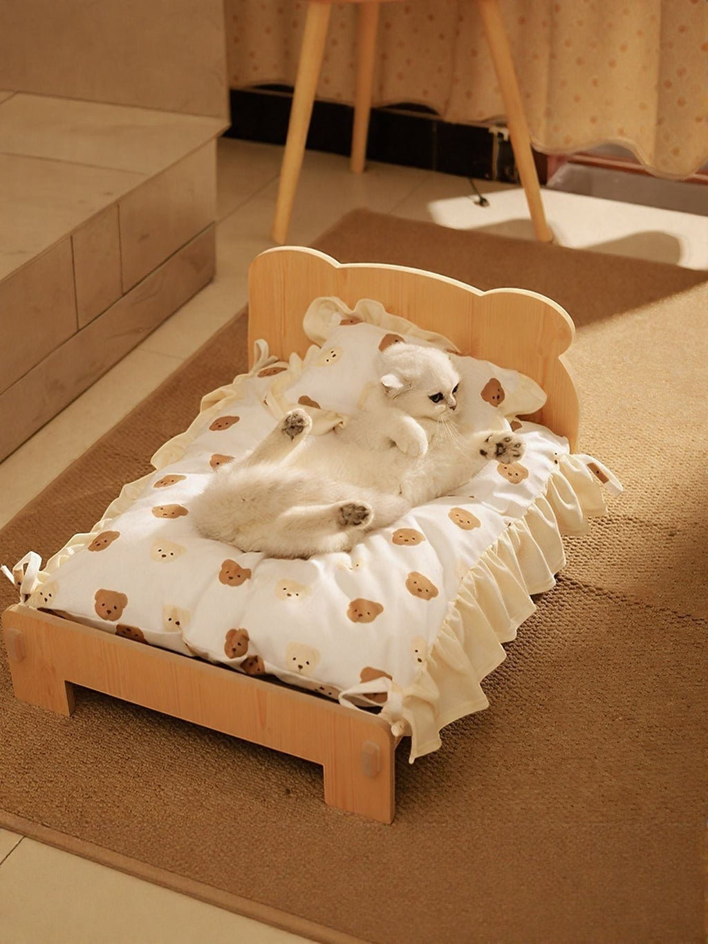 Little Pets Club wooden pet bed with a white cushion featuring a teddy bear print and ruffled edges. A white cat rests on the bed.
