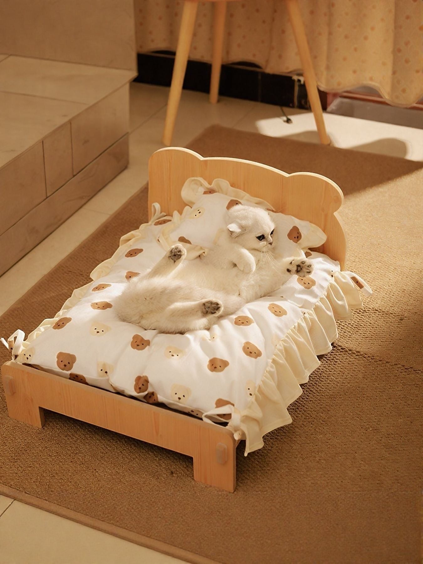 Little Pets Club wooden pet bed with a white cushion featuring a teddy bear print and ruffled edges. A white cat rests on the bed.